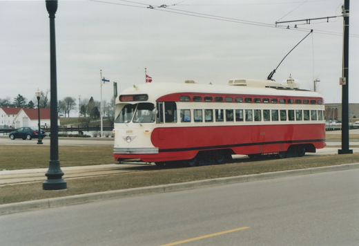 A red and cream streetcar with a trolley pole runs on a grassy median track beside an empty street on an overcast day, with flags, a white building with a red roof and bare trees behind.