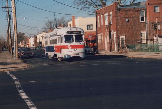 A white streetcar with a red and blue stripe, signed 23 Chestnut Hill, rounds a corner on a street of brick rowhouses under overhead wires, with cars parked along the curbs.