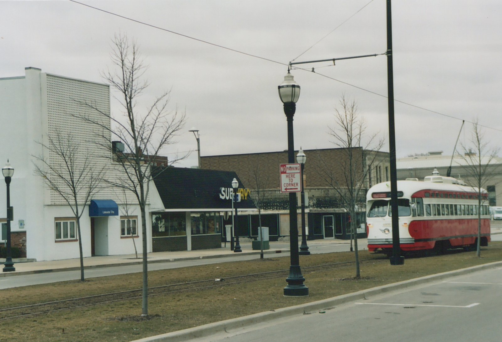 A red and cream streetcar with a trolley pole stands on a track in a grassy median, with bare trees, lamp posts, a No Parking sign and storefronts including a Subway restaurant behind it.