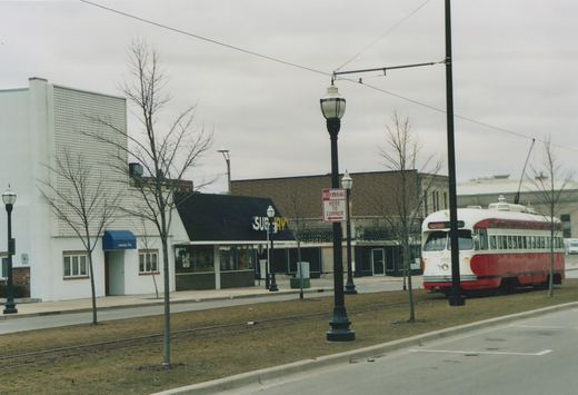 A red and cream streetcar with a trolley pole stands on a track in a grassy median, with bare trees, lamp posts, a No Parking sign and storefronts including a Subway restaurant behind it.