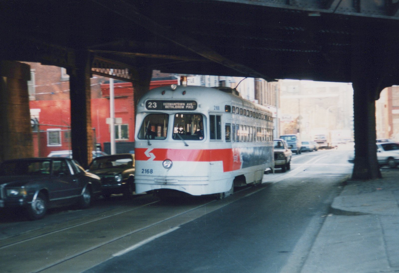 A gray and white streetcar with a red band, signed 23 Germantown Ave Bethlehem Pike, rolls along a street beneath a dark steel viaduct with cars parked beside it.