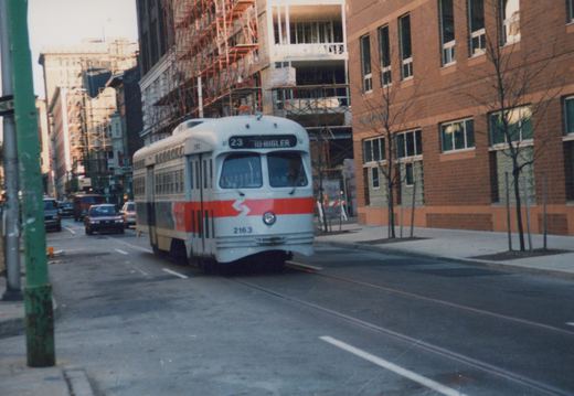A light gray streetcar with a red front band, number 2163, signed 23 TO-BIGLER, runs on street track past a brick building and a construction site with scaffolding.