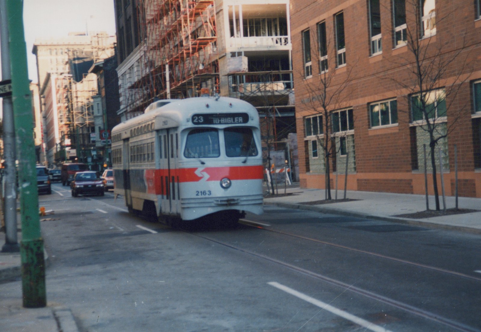 A light gray streetcar with a red front band, number 2163, signed 23 TO-BIGLER, runs on street track past a brick building and a construction site with scaffolding.