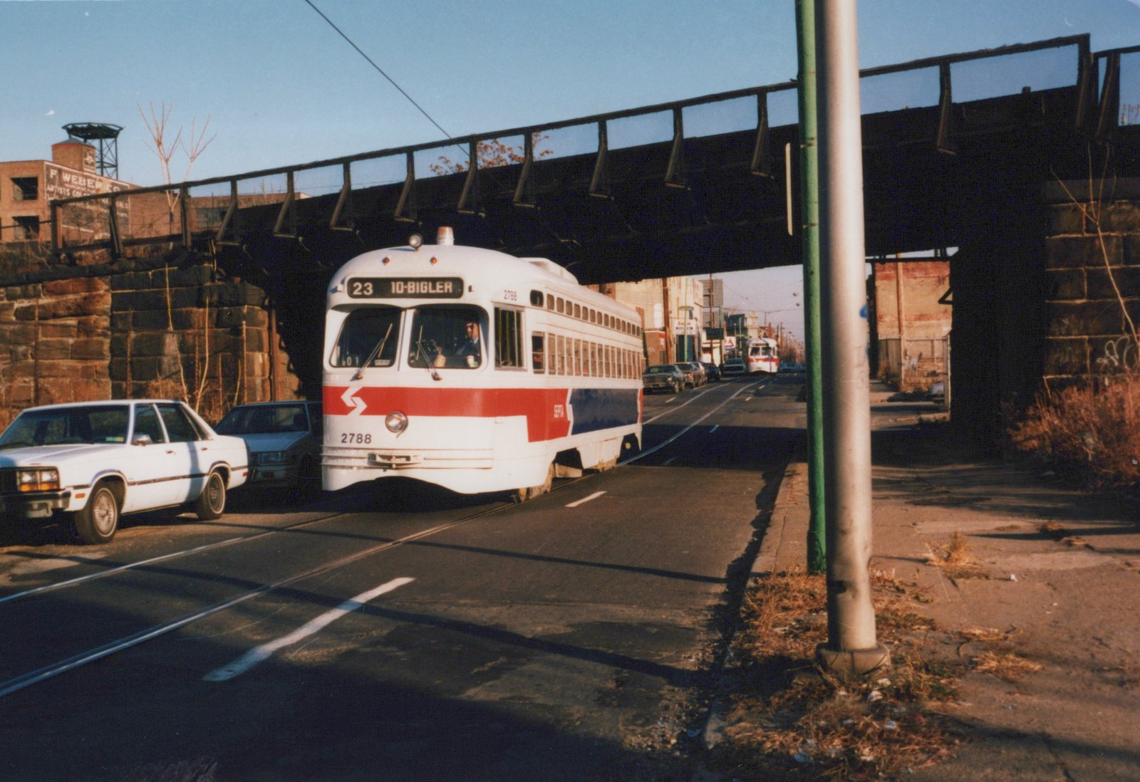 A white streetcar with a red and blue stripe and a 23 10-BIGLER sign passes under a dark steel railroad bridge, with parked cars on the left and a second streetcar in the distance.
