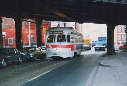 A white streetcar with a red front stripe and a 23 Germantown Ave Bethlehem Pike sign comes toward the camera under a dark steel viaduct, with parked cars on the left and traffic behind.