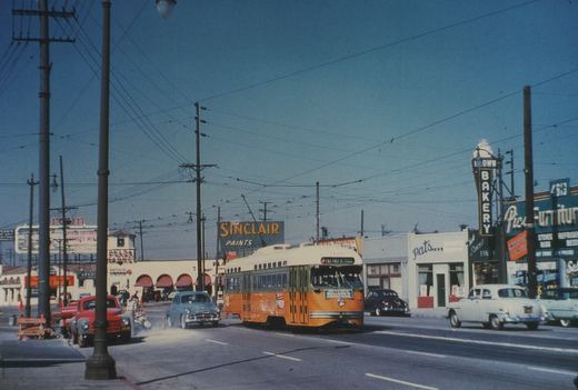 An orange, green and cream streetcar with a trolley pole runs down a sunny street under a web of overhead wires, with a Sinclair Paints billboard, an arcaded market building and small shops, and 1950s cars on the street.
