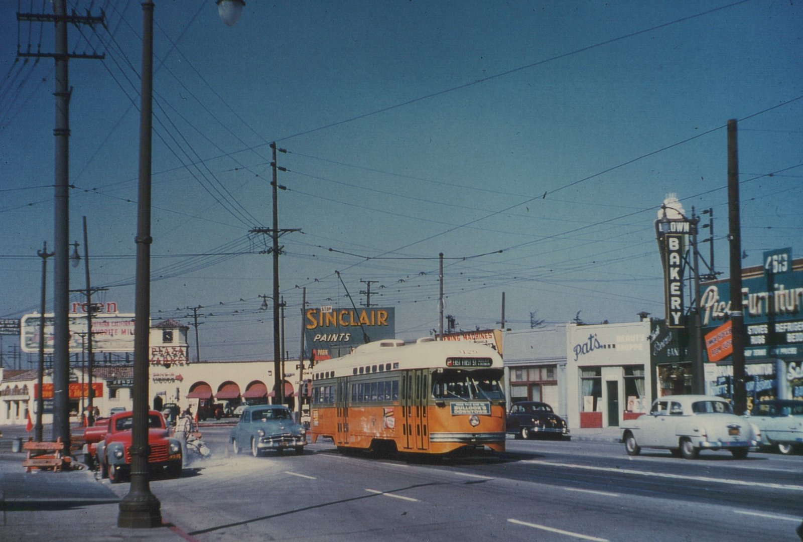An orange, green and cream streetcar with a trolley pole runs down a sunny street under a web of overhead wires, with a Sinclair Paints billboard, an arcaded market building and small shops, and 1950s cars on the street.