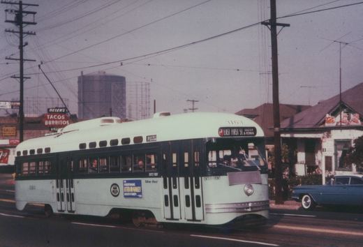 A light green and dark green streetcar with a trolley pole runs along a street beneath overhead wires, with gas holders, a Tacos Burritos sign and a house with Christmas decorations behind it.