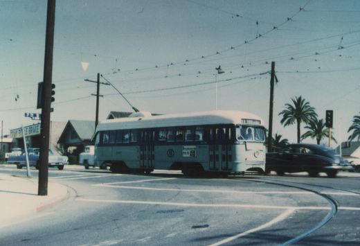 A light green streetcar with a dark window band and cream roof turns through an intersection under a web of overhead wires, with palm trees, a traffic signal and parked cars around it.