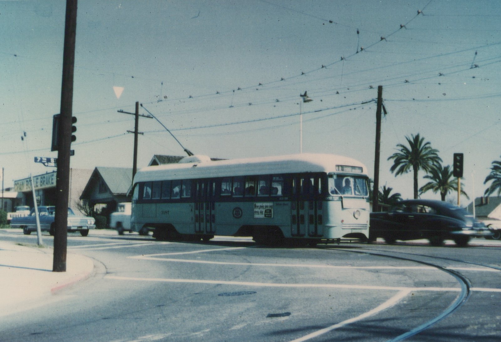 A light green streetcar with a dark window band and cream roof turns through an intersection under a web of overhead wires, with palm trees, a traffic signal and parked cars around it.