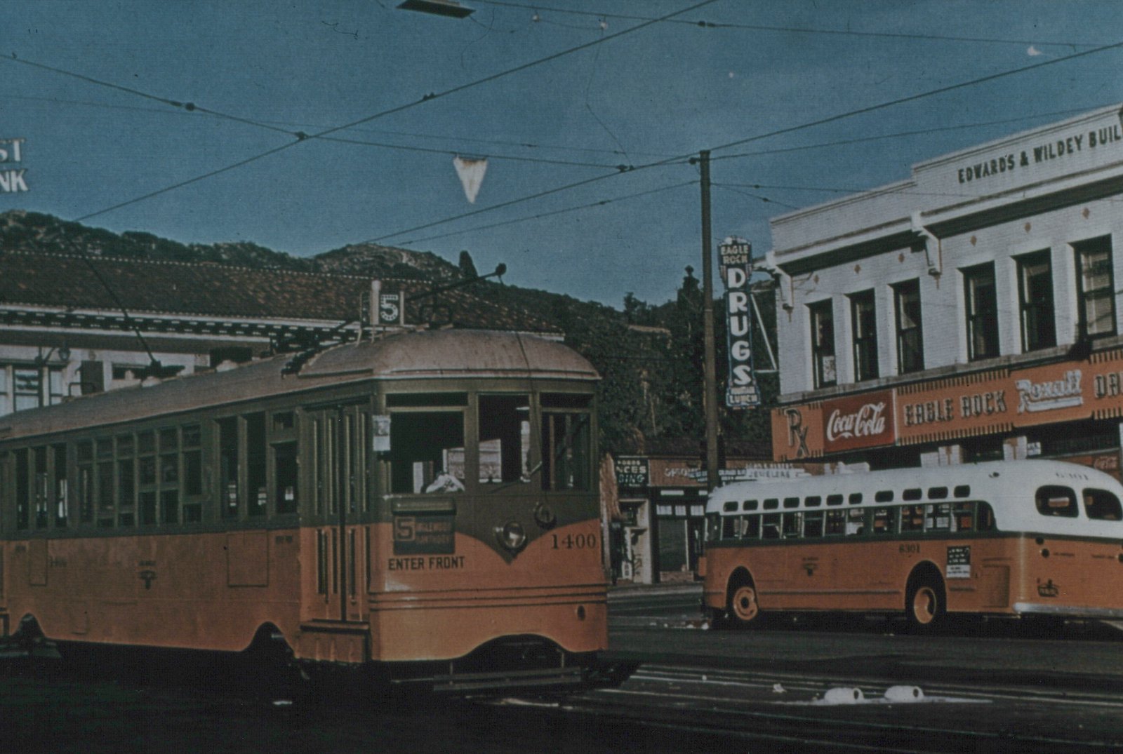 A yellow and green streetcar numbered 1400, with route 5 on its roof sign, stands on a street under trolley wires. A yellow bus with a white roof is parked beside a two-story drugstore building with Coca-Cola and Rexall signs, and hills rise behind.