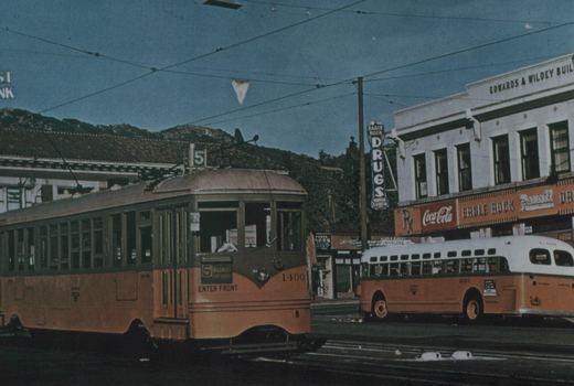 A yellow and green streetcar numbered 1400, with route 5 on its roof sign, stands on a street under trolley wires. A yellow bus with a white roof is parked beside a two-story drugstore building with Coca-Cola and Rexall signs, and hills rise behind.