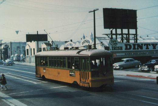 A yellow streetcar with a green window band and a trolley pole crosses an intersection in front of a car lot with a large DINTY sign, while a pedestrian walks across the street at left.