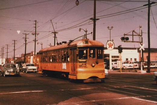 A yellow streetcar with an S route sign and the number 1428 on its dash crosses a railroad track at a street intersection under overhead wires, with a Texaco station and 1950s cars behind it.