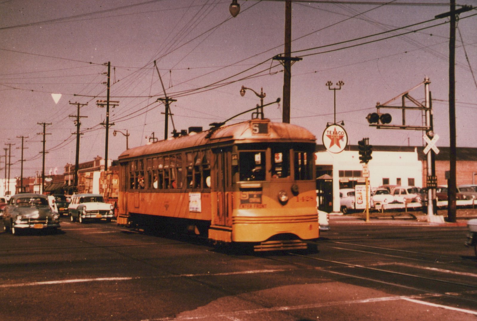 A yellow streetcar with an S route sign and the number 1428 on its dash crosses a railroad track at a street intersection under overhead wires, with a Texaco station and 1950s cars behind it.