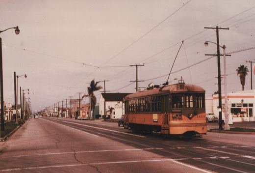 A yellow and green streetcar showing route S and the number 1424 travels on street tracks under trolley wire past a gas station, palm trees and long lines of utility poles on an overcast day.