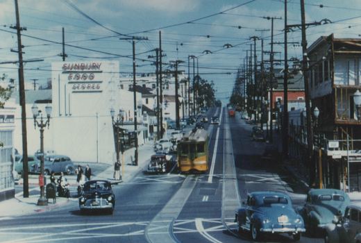 Color photo looking down a long, straight street lined with poles and a dense web of overhead wires. A yellow-and-green streetcar comes up the center tracks toward the camera, with 1940s cars around it and a group of people on the corner at left.
