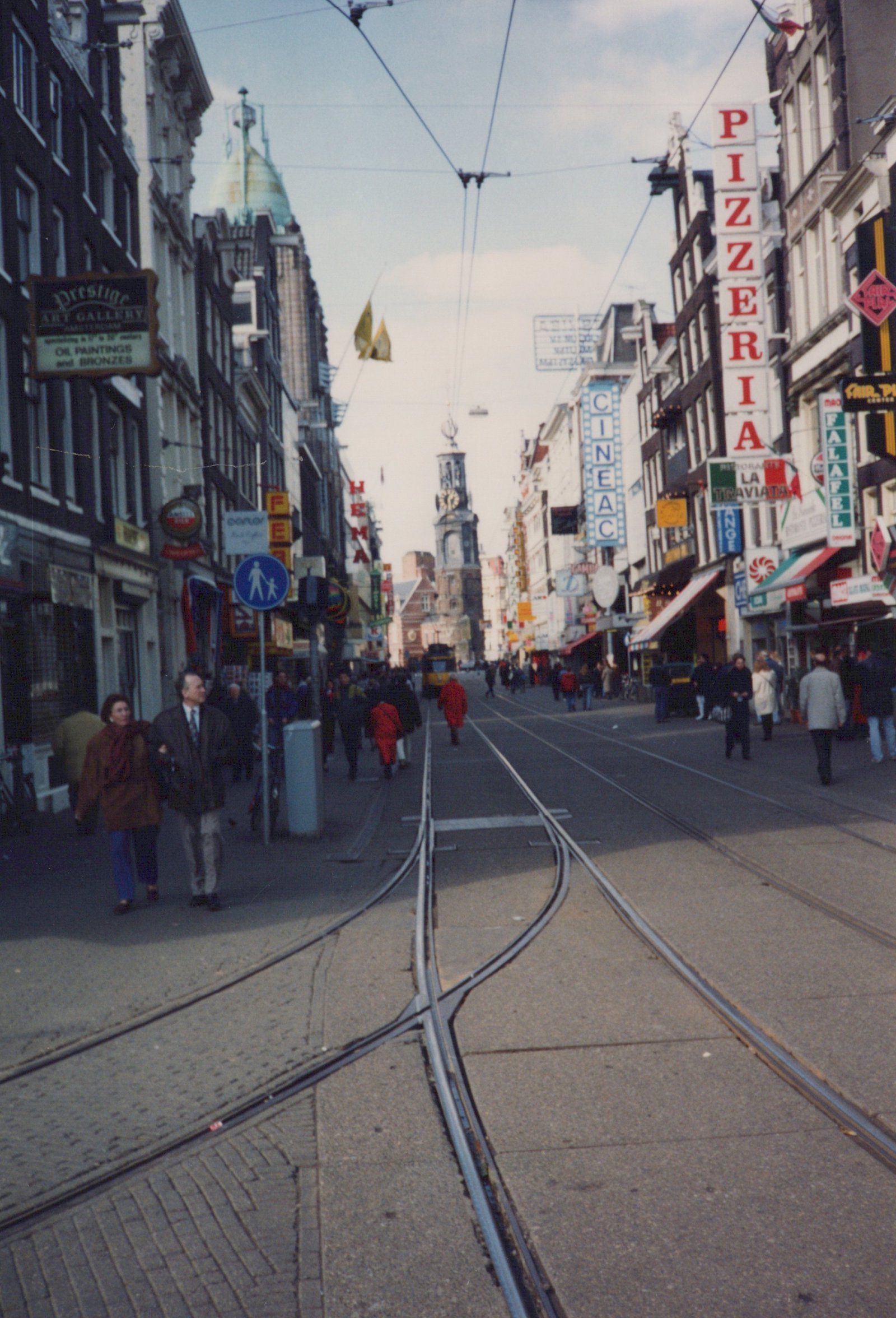 A European city street with tram tracks running down the center, flanked by buildings with signs for a pizzeria and an art gallery, and people walking on the sidewalks.