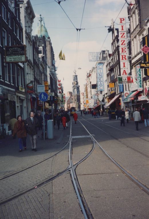 A European city street with tram tracks running down the center, flanked by buildings with signs for a pizzeria and an art gallery, and people walking on the sidewalks.