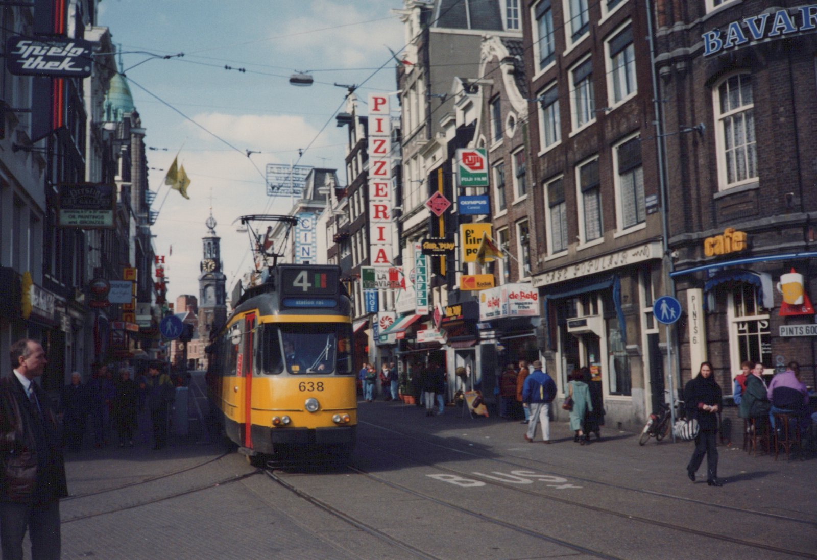 A yellow articulated tram, numbered 638 and signed 4 station rai, comes down a busy shopping street with tram tracks, lined with brick buildings and shop signs, and a clock tower in the distance.