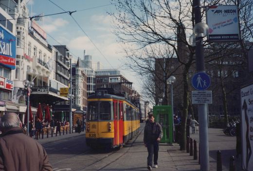 A yellow articulated tram with red doors and route number 4 on its roof comes along a city street lined with cafés and bare trees, while a pedestrian walks toward the camera on the sidewalk.