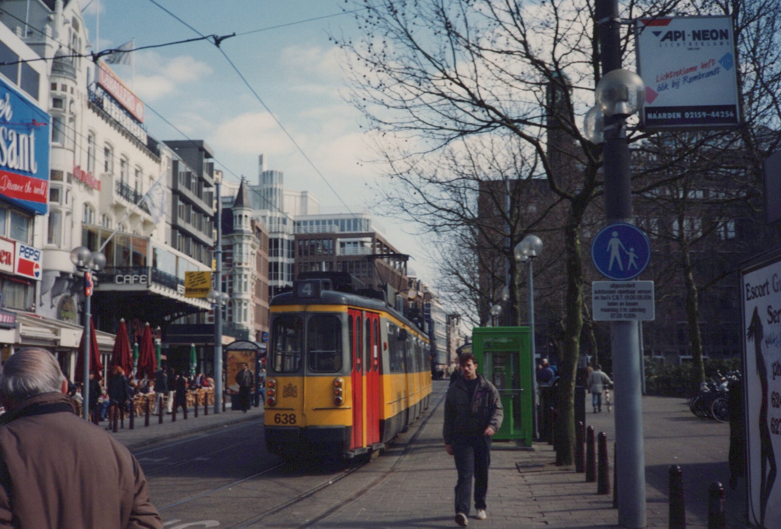 A yellow articulated tram with red doors and route number 4 on its roof comes along a city street lined with cafés and bare trees, while a pedestrian walks toward the camera on the sidewalk.