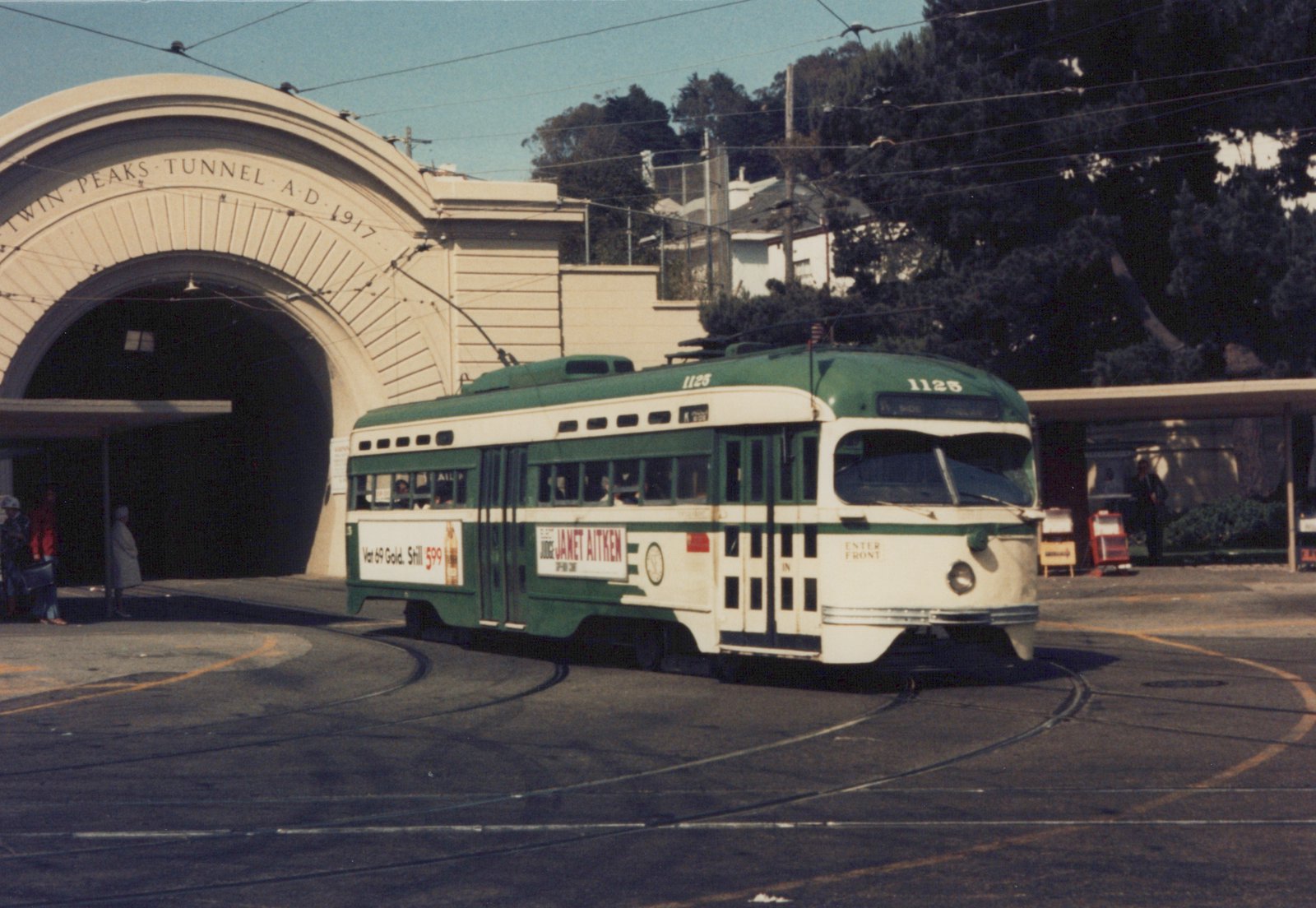 A green and cream streetcar with a trolley pole rounds a curve of track in front of a large arched tunnel portal inscribed Twin Peaks Tunnel A.D. 1917, with people waiting under a shelter.