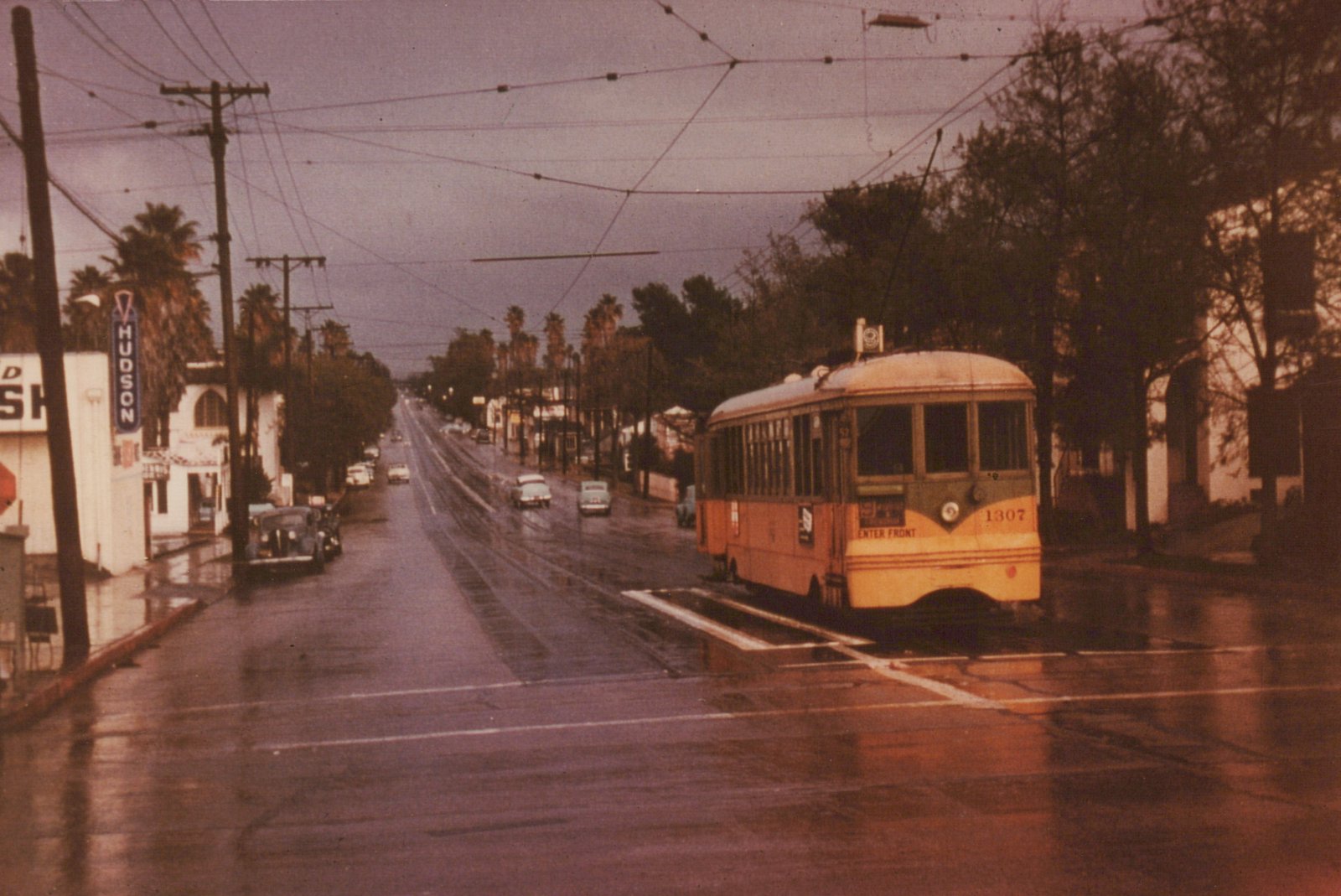 A yellow and green streetcar numbered 1307, with its trolley pole raised, stands beside a raised concrete safety island on a wet street under a stormy sky, with palm trees, a HUDSON sign and 1950s cars lining the road behind it.