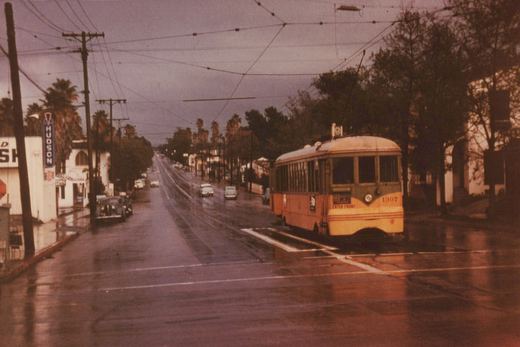 A yellow and green streetcar numbered 1307, with its trolley pole raised, stands beside a raised concrete safety island on a wet street under a stormy sky, with palm trees, a HUDSON sign and 1950s cars lining the road behind it.
