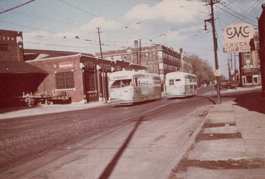 Two light green and cream Chicago Transit Authority PCC streetcars, numbered 4147, run on a street with overhead wires, near a brick building and a GMC Trucks sign, on a sunny day.