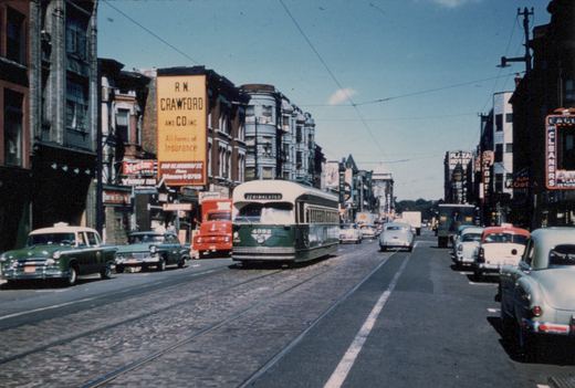 A green and cream PCC streetcar with the number 4382 and destination sign '22 HALSTED' is shown on a sunlit, cobblestone city street, surrounded by 1950s cars and storefronts under a clear blue sky.