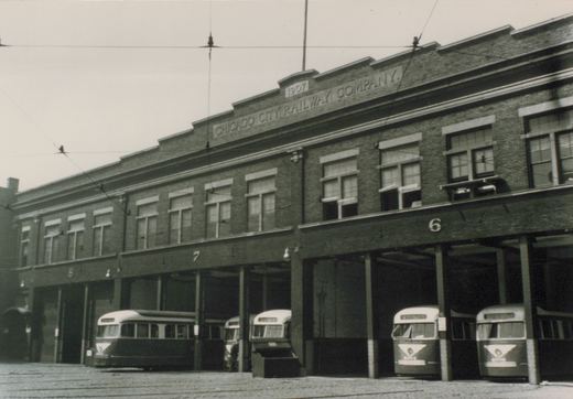 A black-and-white photo of a long two-story brick carbarn lettered CHICAGO CITY RAILWAY COMPANY and dated 1907, with five streamlined two-tone streetcars nosed out of its numbered bays under trolley wires.