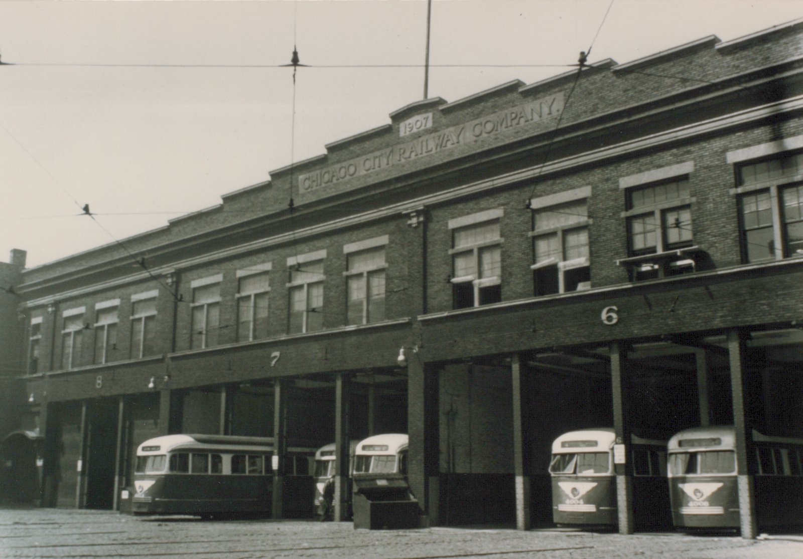 A black-and-white photo of a long two-story brick carbarn lettered CHICAGO CITY RAILWAY COMPANY and dated 1907, with five streamlined two-tone streetcars nosed out of its numbered bays under trolley wires.