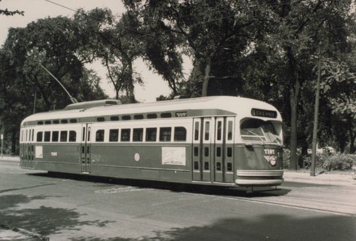 Black-and-white photo of a two-tone streamlined streetcar with its trolley pole raised, signed 36 STATE-KINZIE, on a street under tall trees.