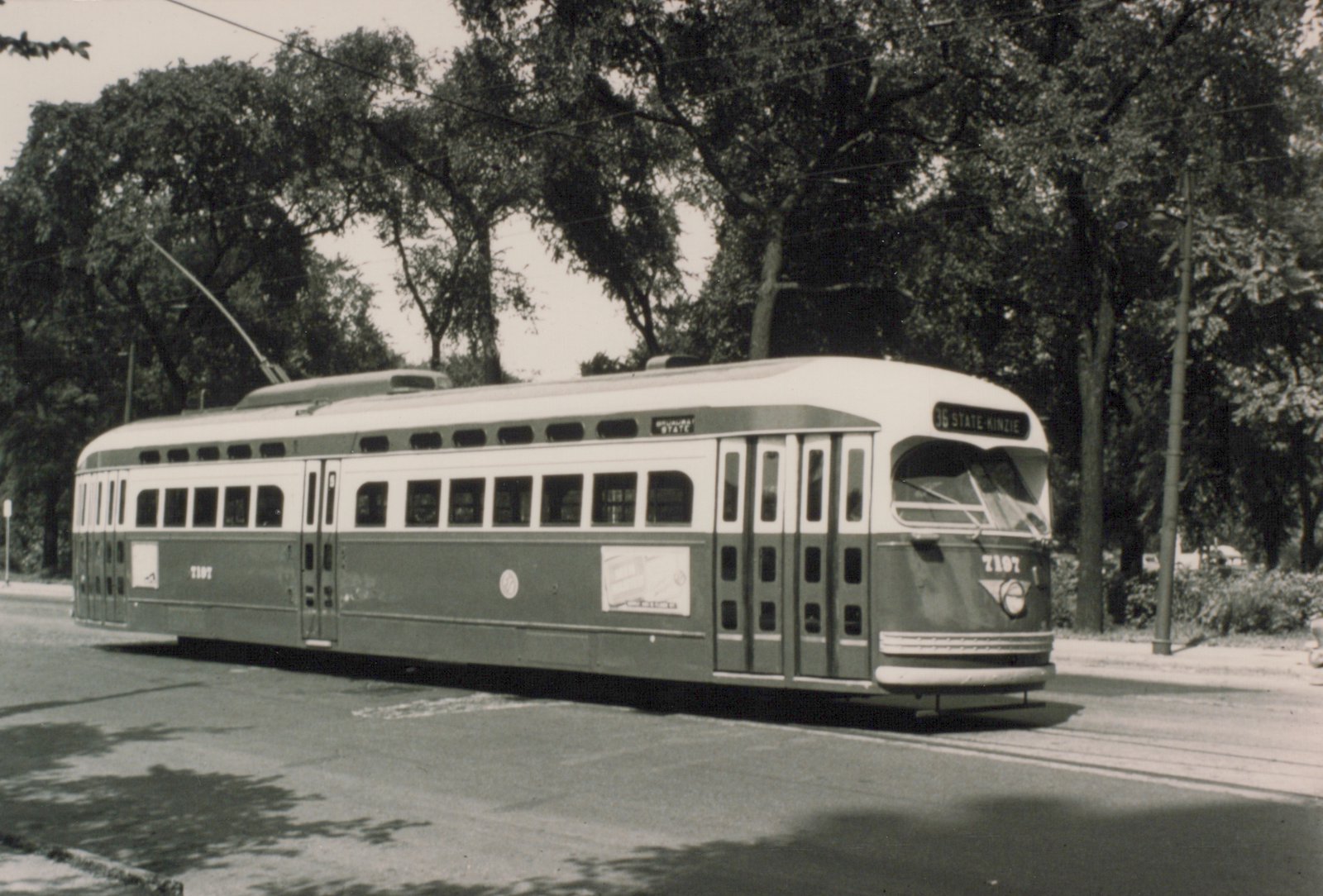 Black-and-white photo of a two-tone streamlined streetcar with its trolley pole raised, signed 36 STATE-KINZIE, on a street under tall trees.