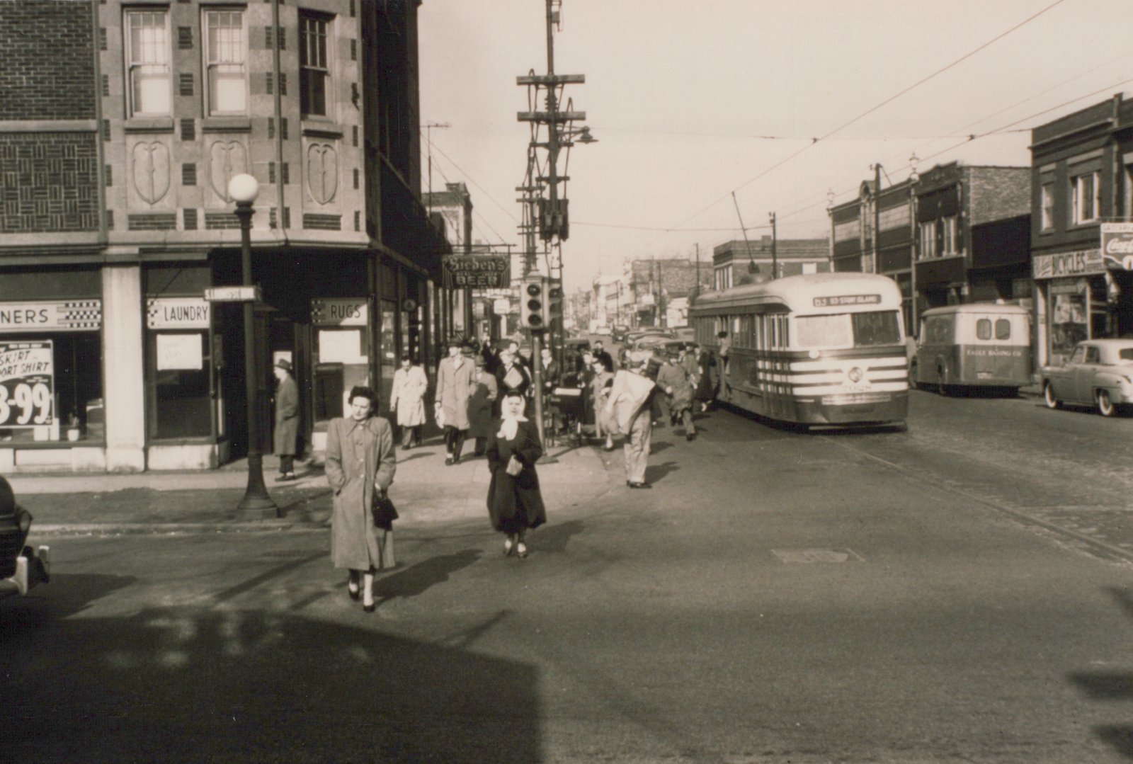 Sepia street scene with a streamlined streetcar under trolley wires on a shop-lined street, while pedestrians cross the intersection and a crowd stands on the corner.