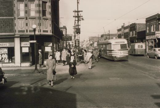 Sepia street scene with a streamlined streetcar under trolley wires on a shop-lined street, while pedestrians cross the intersection and a crowd stands on the corner.
