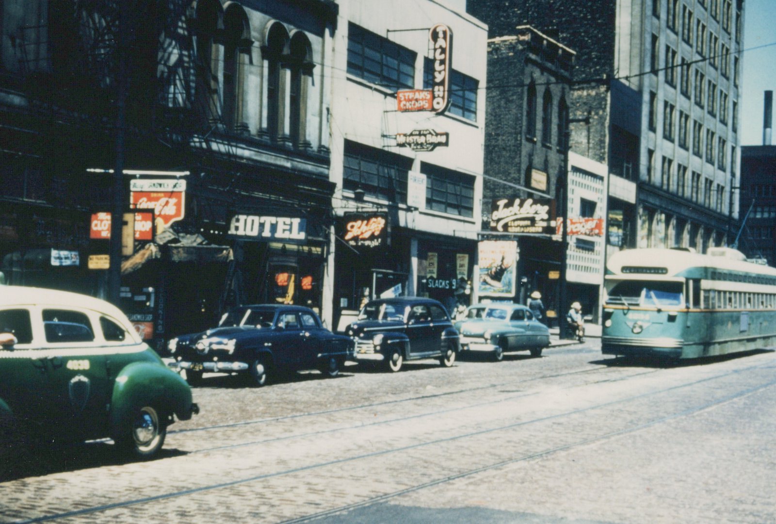 A green and cream streetcar comes down a cobbled street with rails, past parked 1940s and 1950s cars and storefronts with Hotel, Tally Ho and Schlitz signs, with a green and cream taxicab in the foreground.