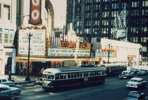 A green and cream streetcar on a city street in front of a brightly lit theatre marquee advertising Run for Cover, with a bus and 1950s cars behind it.