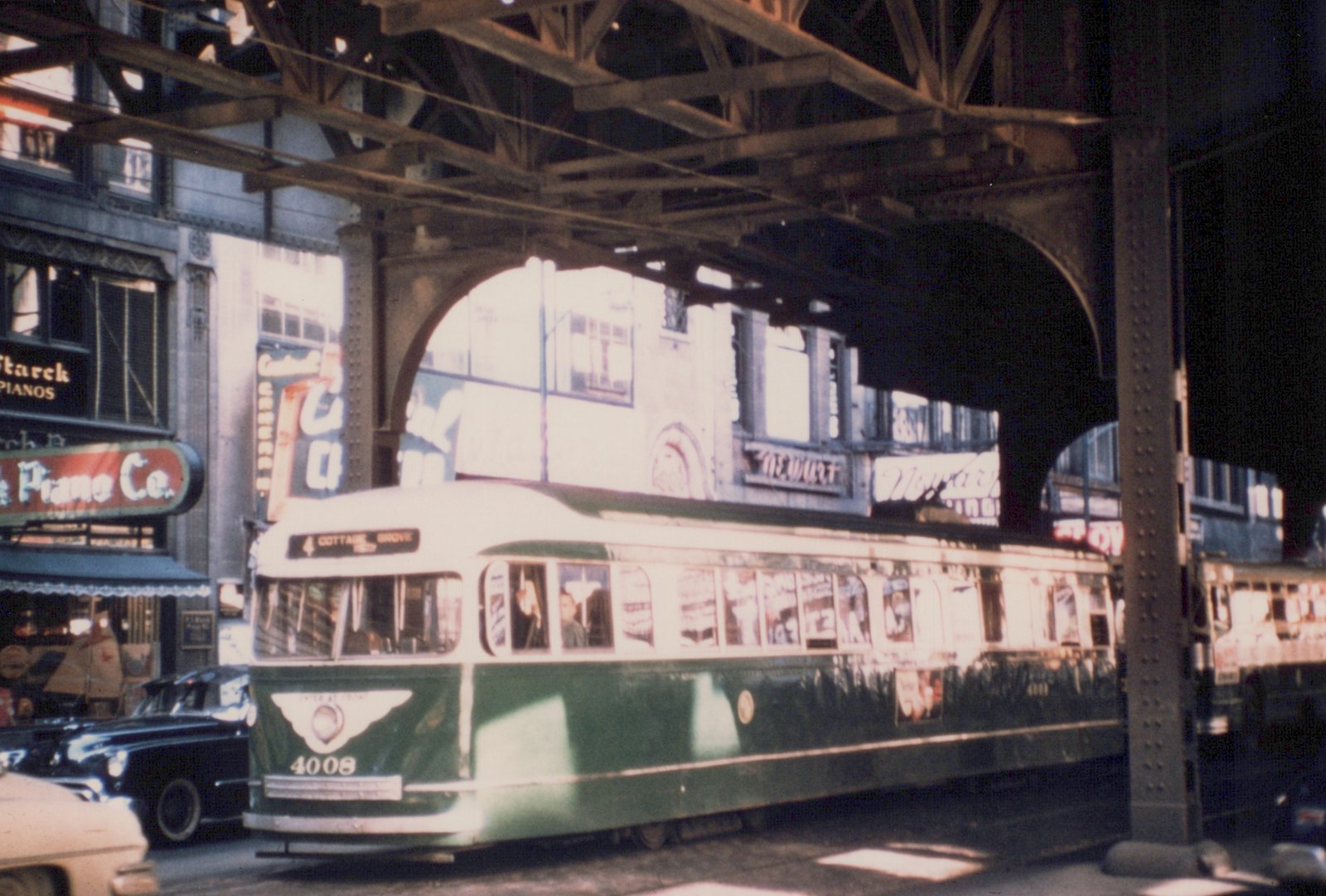 A green and cream streetcar numbered 4008 with a sign reading 4 Cottage Grove runs along a street in the shadow of a steel elevated railway, with piano store signs on the buildings at left.
