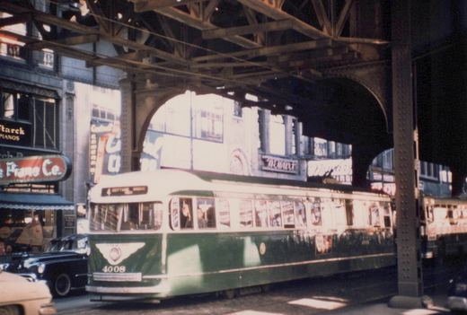 A green and cream streetcar numbered 4008 with a sign reading 4 Cottage Grove runs along a street in the shadow of a steel elevated railway, with piano store signs on the buildings at left.