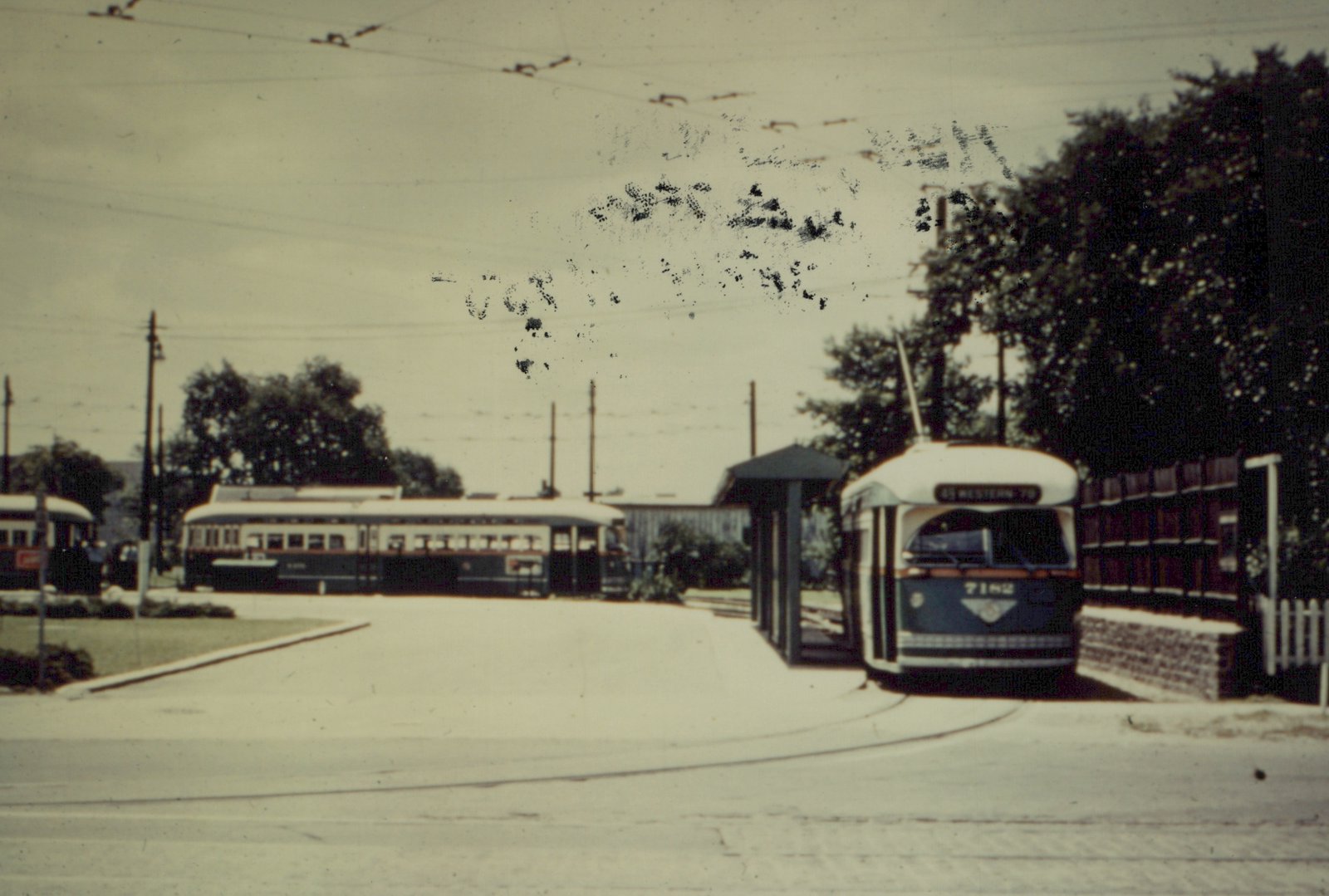 A green and cream streetcar with its trolley pole raised sits on a curved track beside a covered shelter and a wooden fence. A longer, older streetcar and part of another car stand across a paved loop under overhead wires.