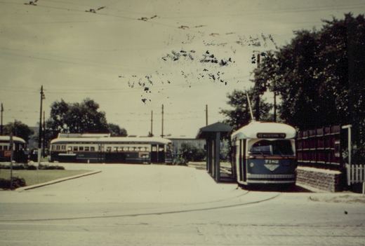 A green and cream streetcar with its trolley pole raised sits on a curved track beside a covered shelter and a wooden fence. A longer, older streetcar and part of another car stand across a paved loop under overhead wires.