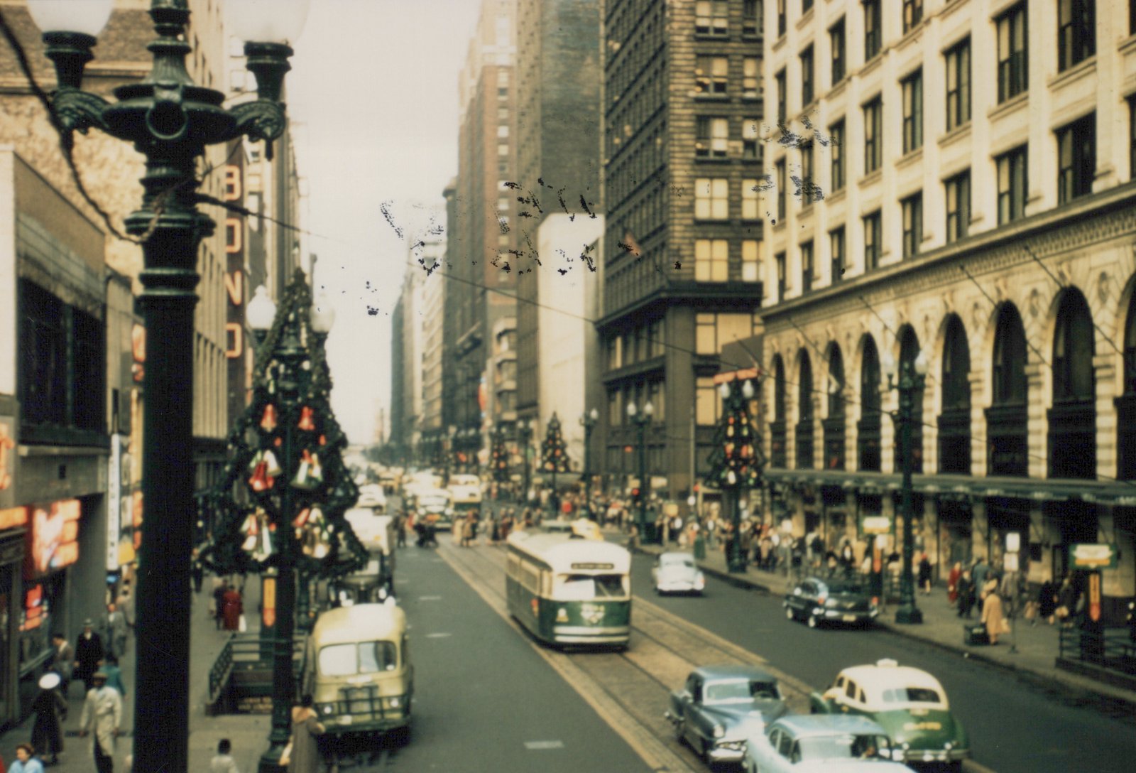 A high view down a busy downtown street decorated for Christmas, with a green and cream streetcar in the center lanes, a bus at the left curb, cars and a taxi in front, and crowds on both sidewalks.