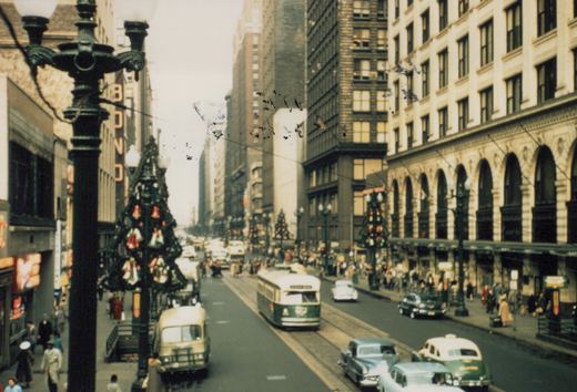 A high view down a busy downtown street decorated for Christmas, with a green and cream streetcar in the center lanes, a bus at the left curb, cars and a taxi in front, and crowds on both sidewalks.