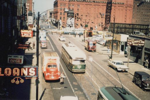 A green and cream streetcar seen from above runs on cobblestone tracks between a red Budweiser truck and parked cars, with neon store signs on the left and a large red brick warehouse in the background.