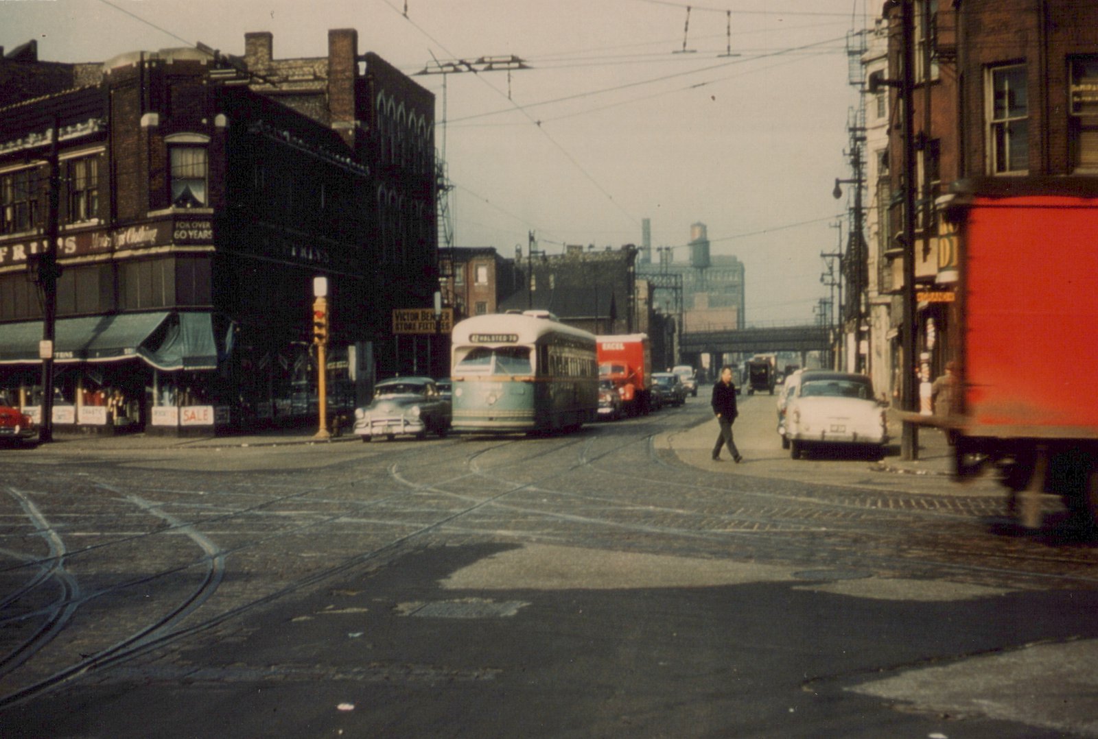 A green and cream streetcar signed 42 HALSTED-79 comes toward the camera under overhead wires. Old brick storefronts line the street, a man walks across the foreground, and a red truck blurs past at the right.