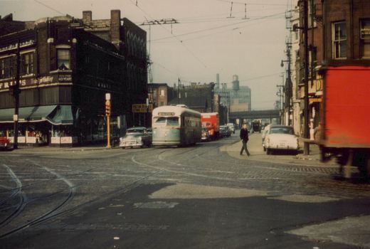 A green and cream streetcar signed 42 HALSTED-79 comes toward the camera under overhead wires. Old brick storefronts line the street, a man walks across the foreground, and a red truck blurs past at the right.