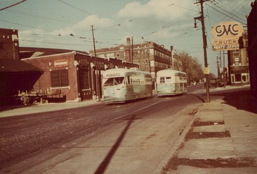 Two green and cream streetcars, one approaching and one going away, on a brick street under trolley wires, with brick warehouses at left and a GMC Trucks sign at right.