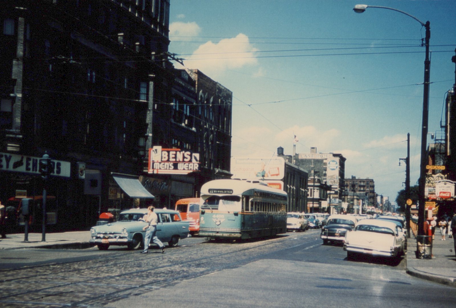 A green and cream streetcar on a cobbled street with tracks under overhead wires. A pale blue station wagon and a man crossing the street are in the foreground, and parked cars and old brick buildings with shop signs line the street.